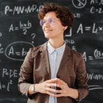 Smiling female teacher standing in front of a mathematical blackboard, illustrating complex equations and teaching concepts.